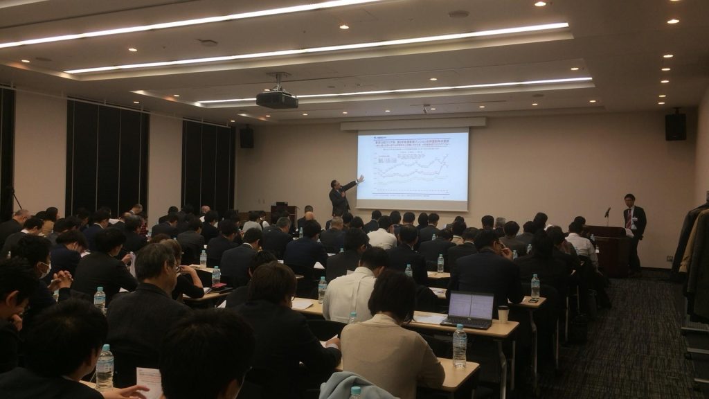 会議室で、大勢の人々が集まり、スクリーンを使用して説明している弊社上席主任研究員である井出武の話を座って聞いている様子を写した写真