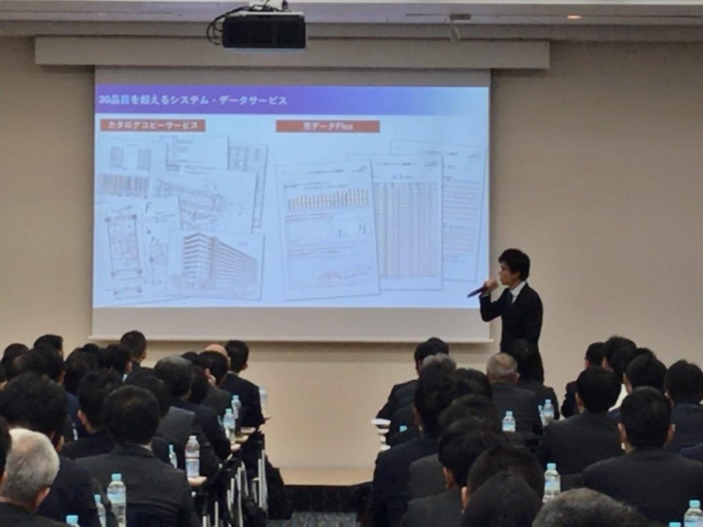 スクリーンの前でプレゼンテーションを行う東京カンテイ不動産営業部課長　髙木大輔と、その話を座って聞いている人々が集まっている様子の写真